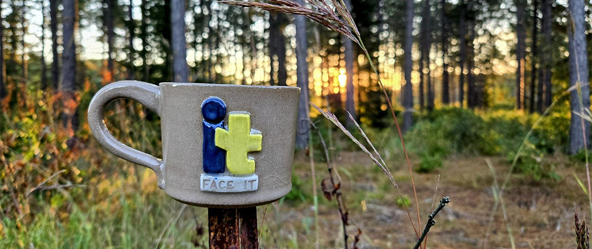 A Face It Foundation mug sitting on a fence post in the woods.